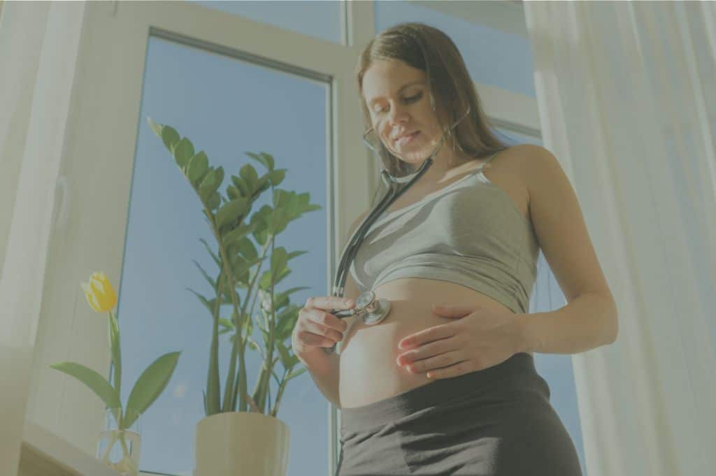 Pregnant woman stands by a window and listens to her baby's heartbeat with a stethescope